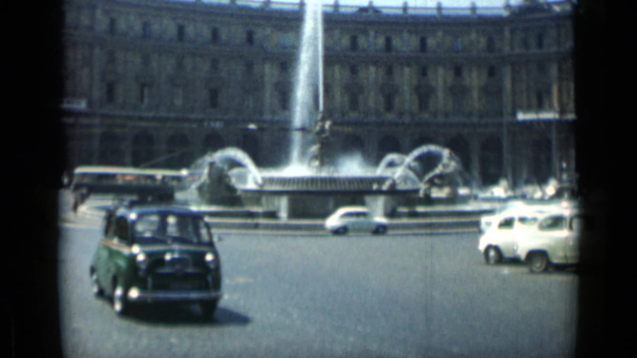 Cars pass around a fountain with a bus and large building in the background.