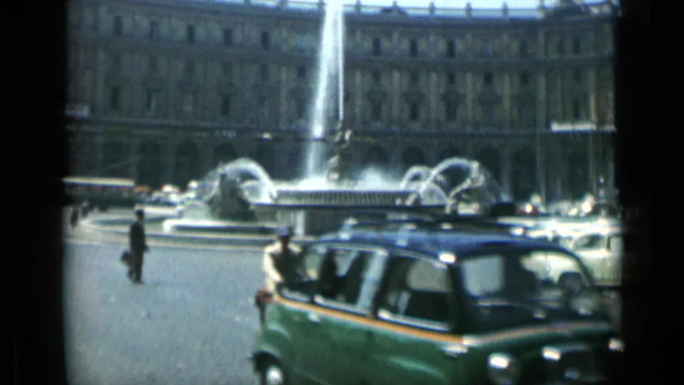 A green mid-century car passes close to the camera as pedestrians and fountain traffic continue in 1962 Rome.
