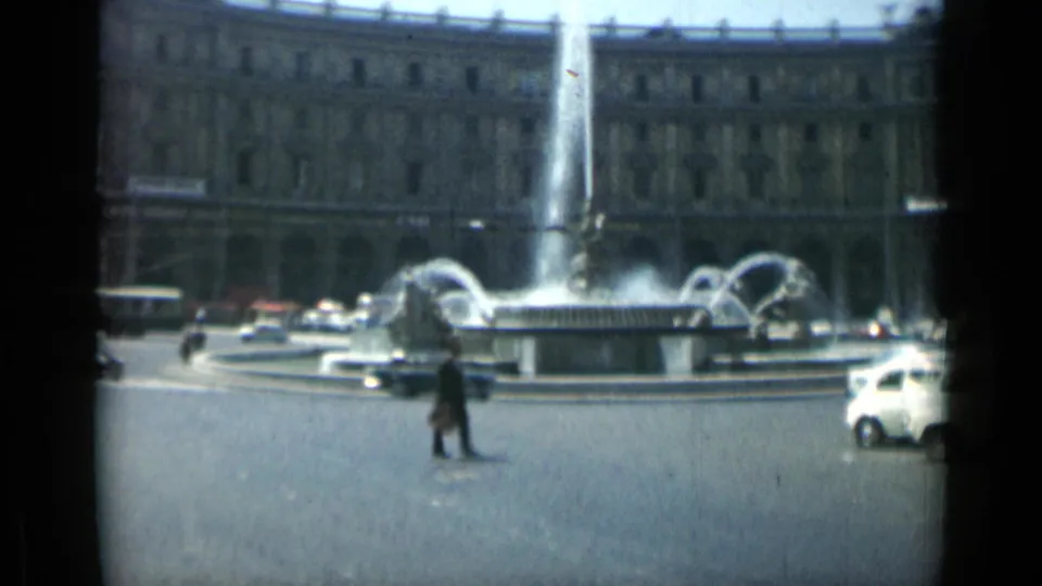 A lone pedestrian crosses the Rome traffic circle as fountain jets rise behind passing cars and a white vehicle.