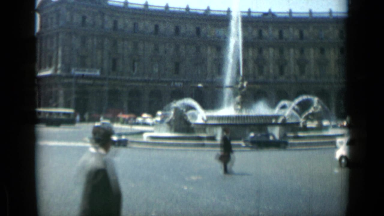 Fountain in a Rome traffic circle with pedestrians, cars, and a large classical building behind it.