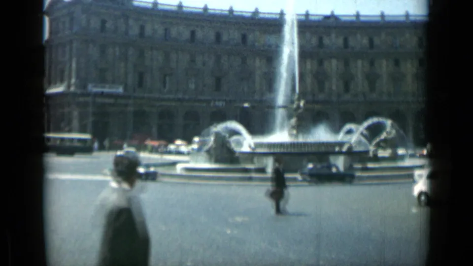 1962 Rome home-movie frame showing a tall fountain spray, crossing pedestrians, period cars, and an ornate building facade.
