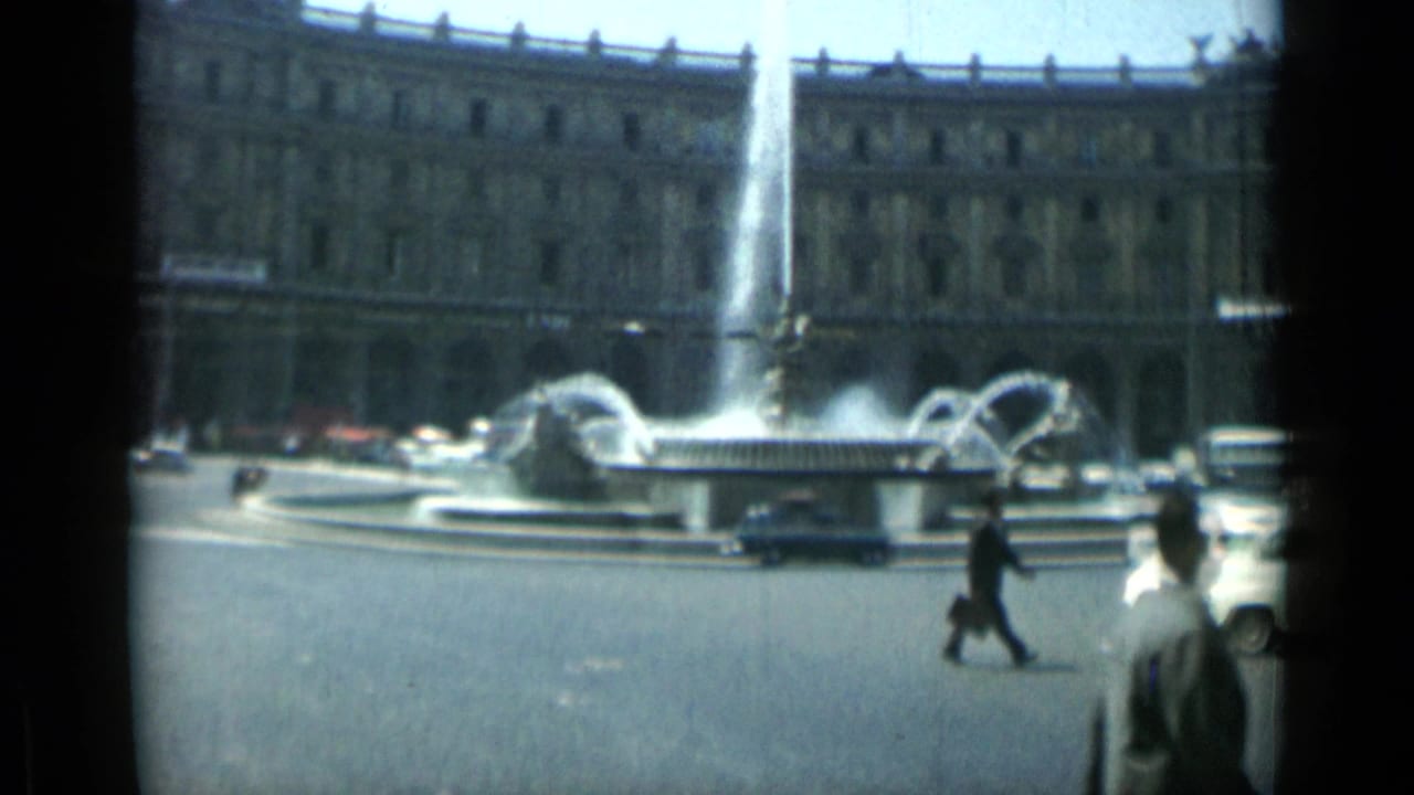 Rome street scene from 1962 with a central fountain, moving cars, a suited pedestrian, and a monumental stone facade.