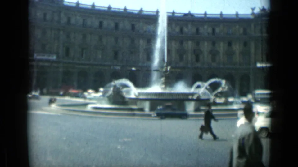 Rome street scene from 1962 with a central fountain, moving cars, a suited pedestrian, and a monumental stone facade.