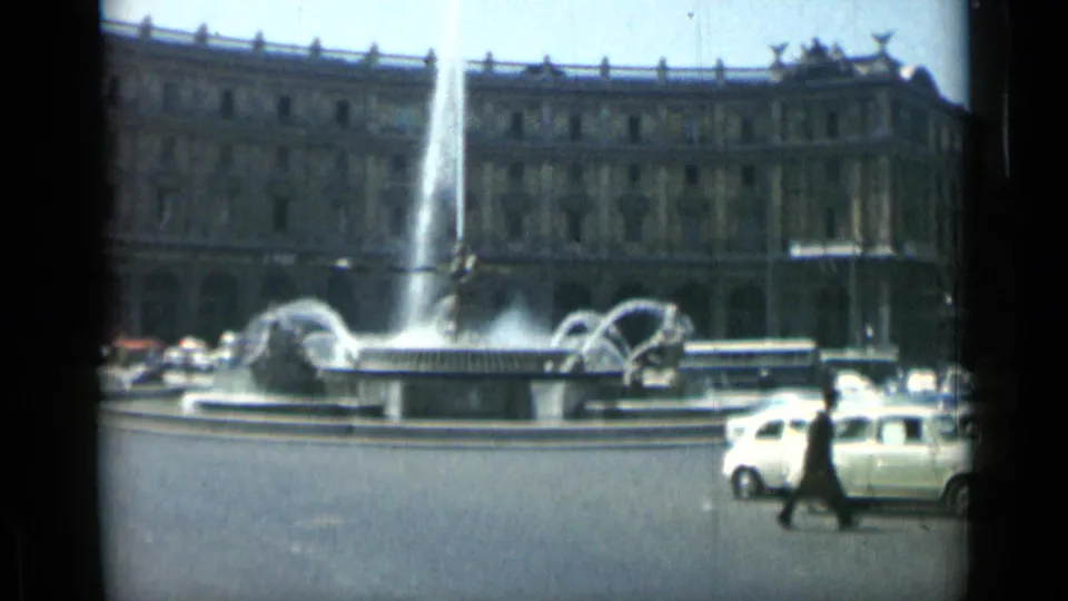 1962 Rome frame with fountain jets, a pale compact car, pedestrians, and the surrounding urban square in bright daylight.