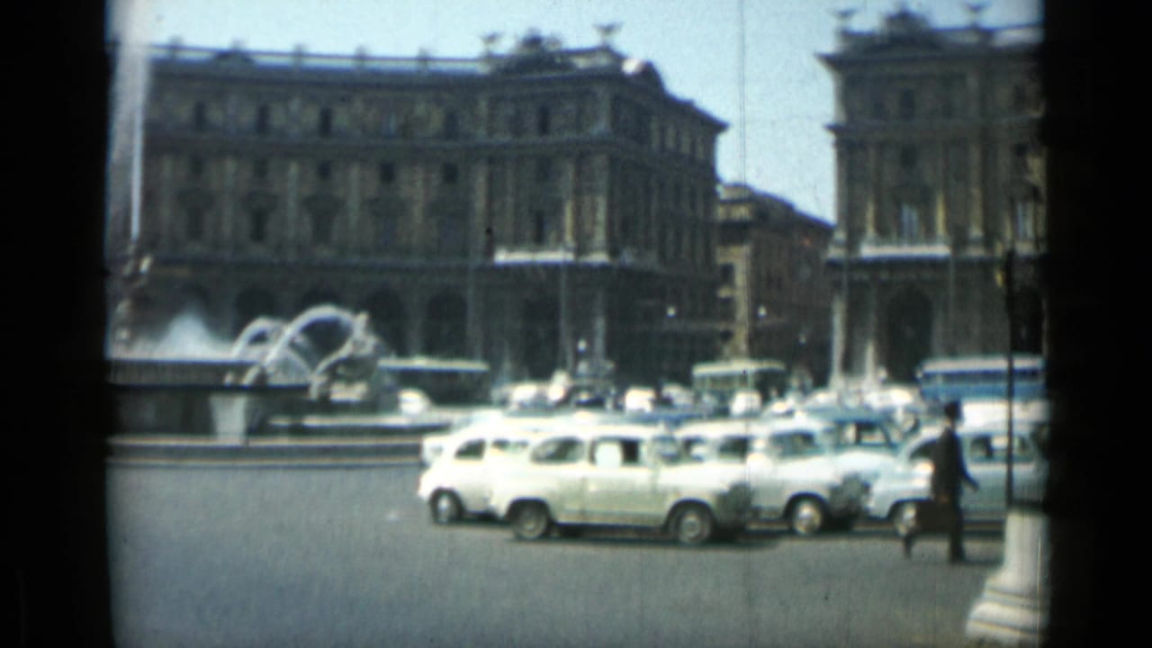 Public square with a grand building, fountain spray, cars, and pedestrians.