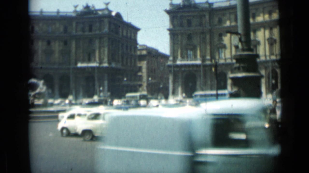 1962 Rome square with light-colored cars and a blurred vehicle passing in front of a public building and fountain.