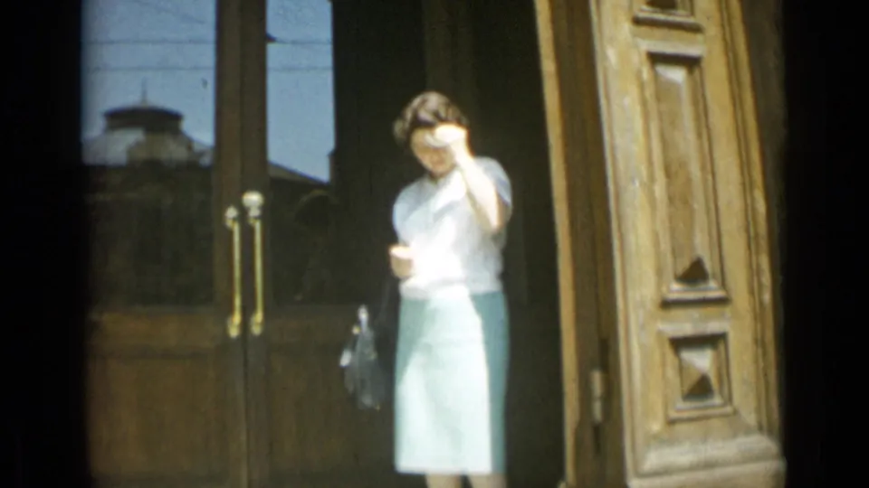 Woman standing at a large doorway in 1962 Rome, wearing a pale blouse and green skirt and holding a small bag.