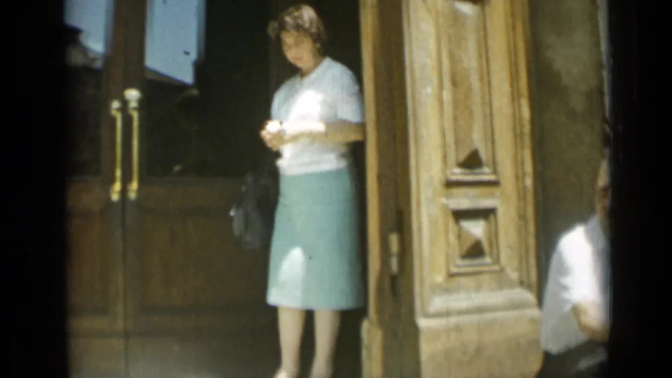 1962 Rome doorway view with a woman in a pale blouse and green skirt, plus a partial figure at the right edge.