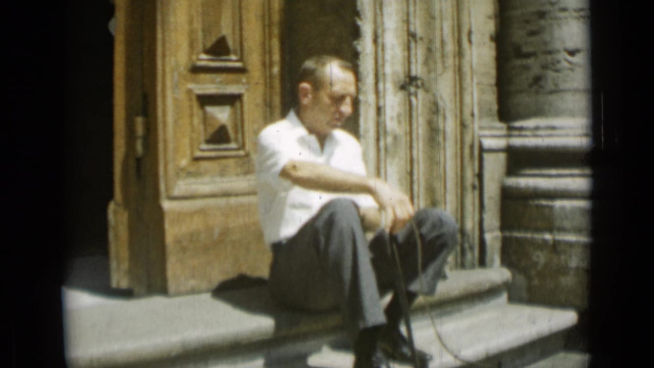 In the 1962 Rome home-movie footage, the man sits near carved wooden doors with his arms resting over one knee.