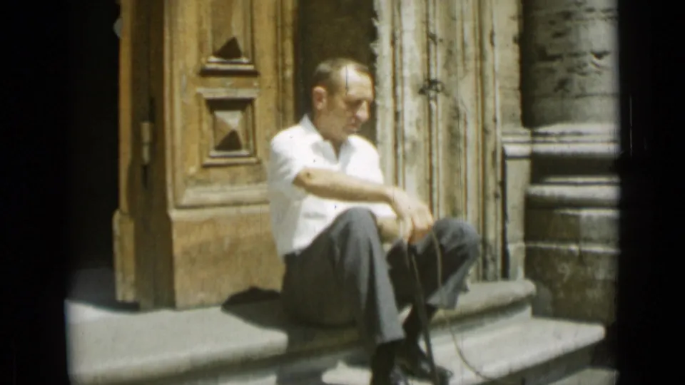 In the 1962 Rome home-movie footage, the man sits near carved wooden doors with his arms resting over one knee.
