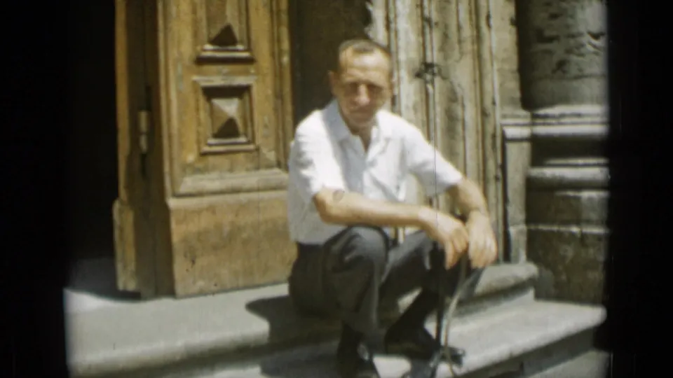 At the end of the Rome 1962 clip, the man faces toward the camera while seated near the carved wooden door and stone column.