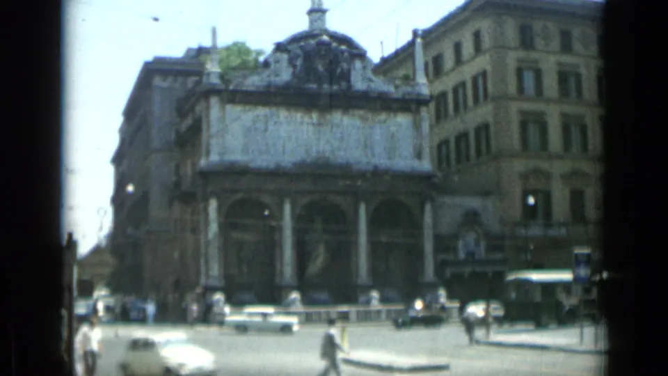 Rome street traffic and pedestrians pass an older columned building in 1962 8mm travel footage.