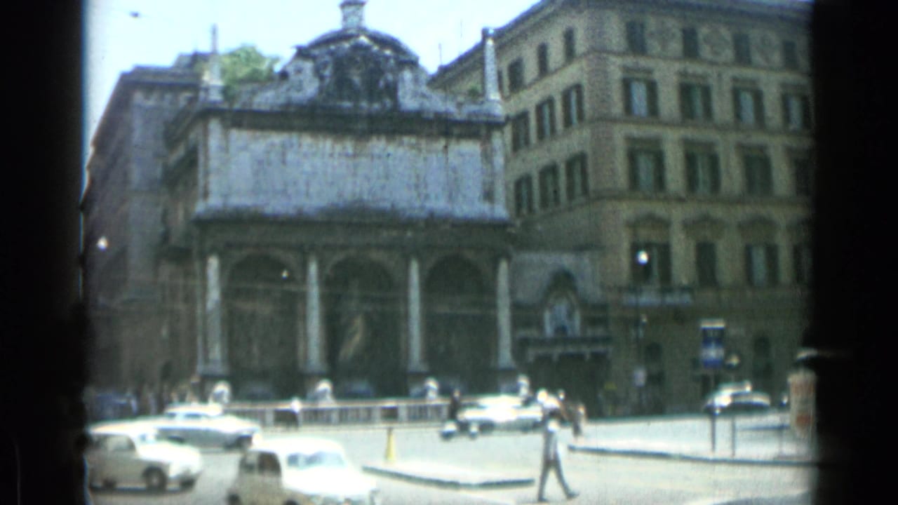 Columned urban building beside a Rome street with cars and pedestrians.