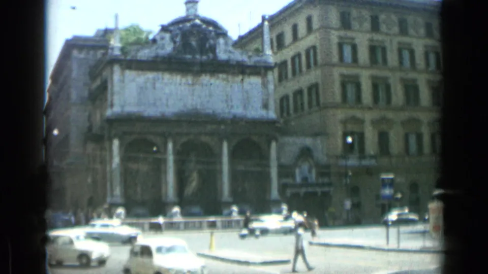 1962 8mm view of a Rome street with a columned building, parked and moving cars, and pedestrians crossing nearby.