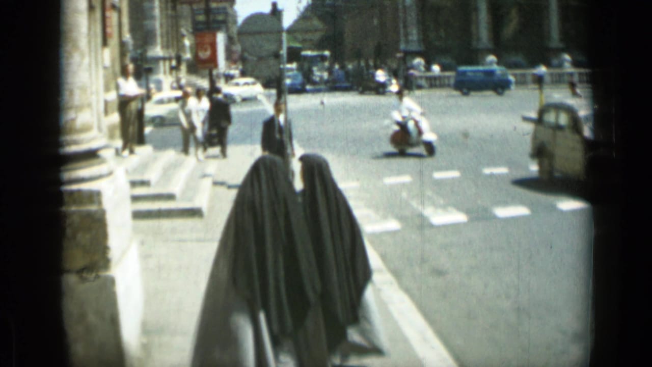 Two nuns walk along a sidewalk beside traffic and pedestrians.