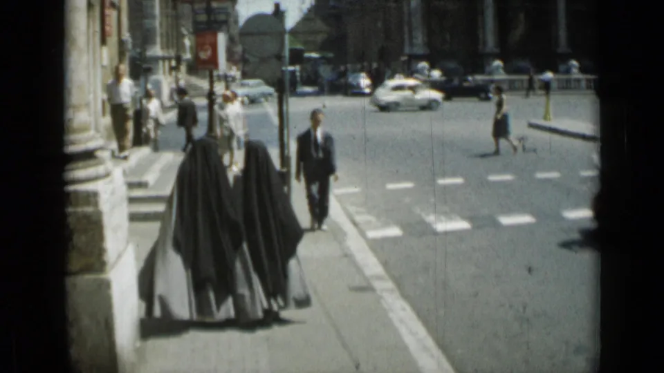 Two nuns in dark habits are filmed from behind on a Rome sidewalk, with suited pedestrians, street markings, and cars in the background.