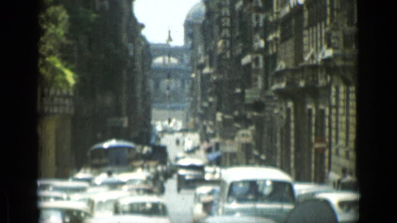 Sunlit Rome street with vintage cars moving between tall buildings.