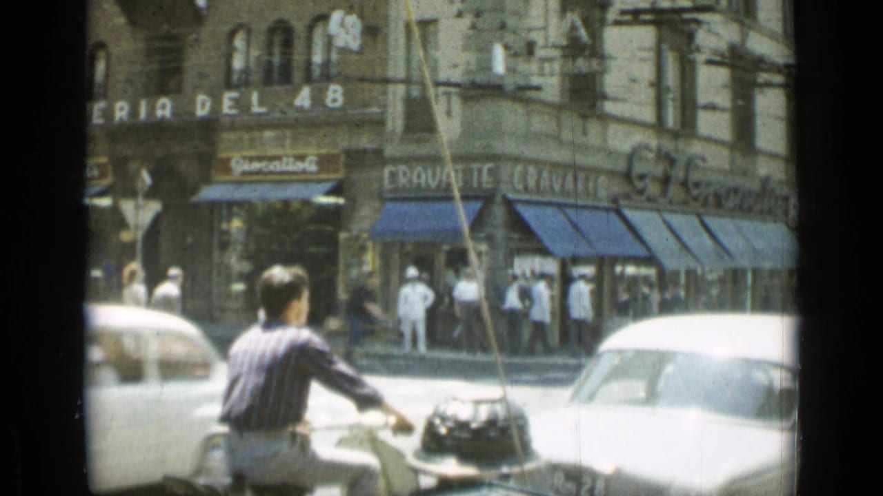 Moped rider passes an intersection near storefronts with blue awnings.