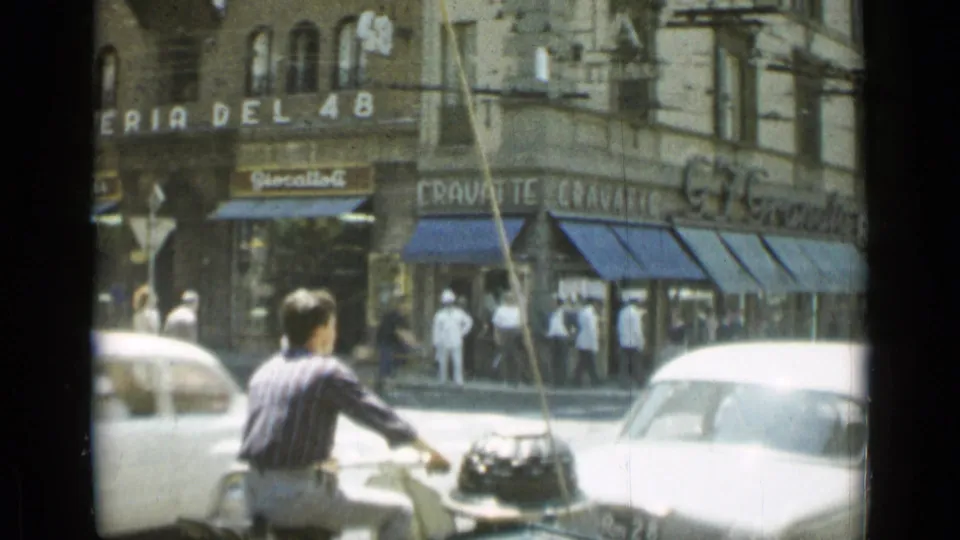 1962 Rome intersection with a moped rider, pedestrians, vintage cars, and storefront signage including a cravatte shop.