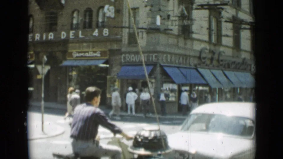 Mid-century Rome street corner with a moped rider, white car, pedestrians, blue awnings, and Italian storefront lettering.