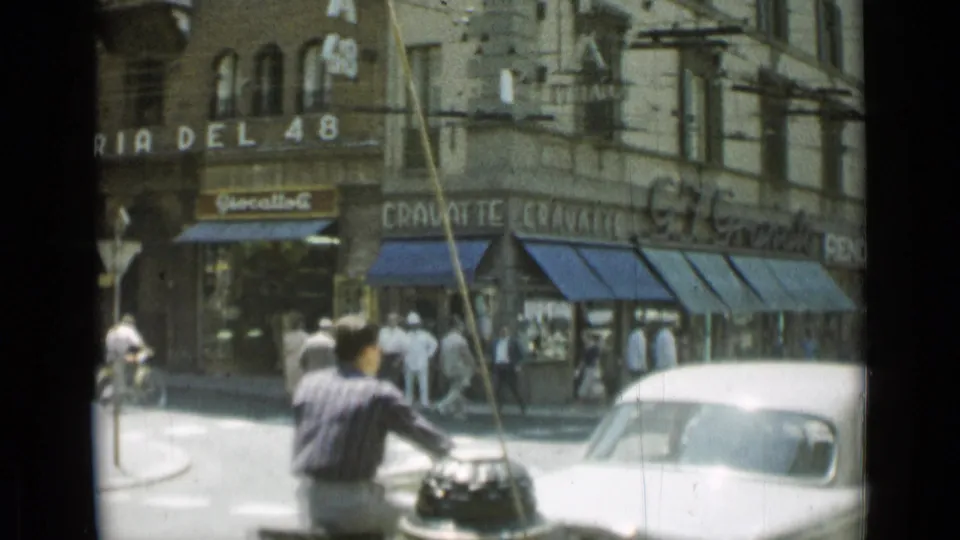 1962 Rome street corner showing pedestrians outside shops, a moped rider in the foreground, and a white car beside blue awnings.