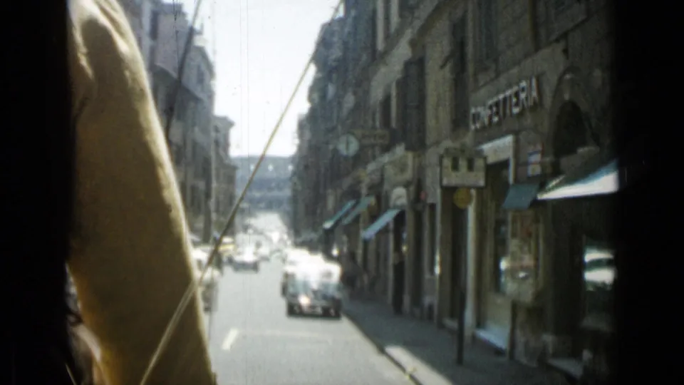 1962 Rome street view from inside a moving vehicle, showing a narrow roadway, cars ahead, stone facades, shop awnings, and a dark interior edge.