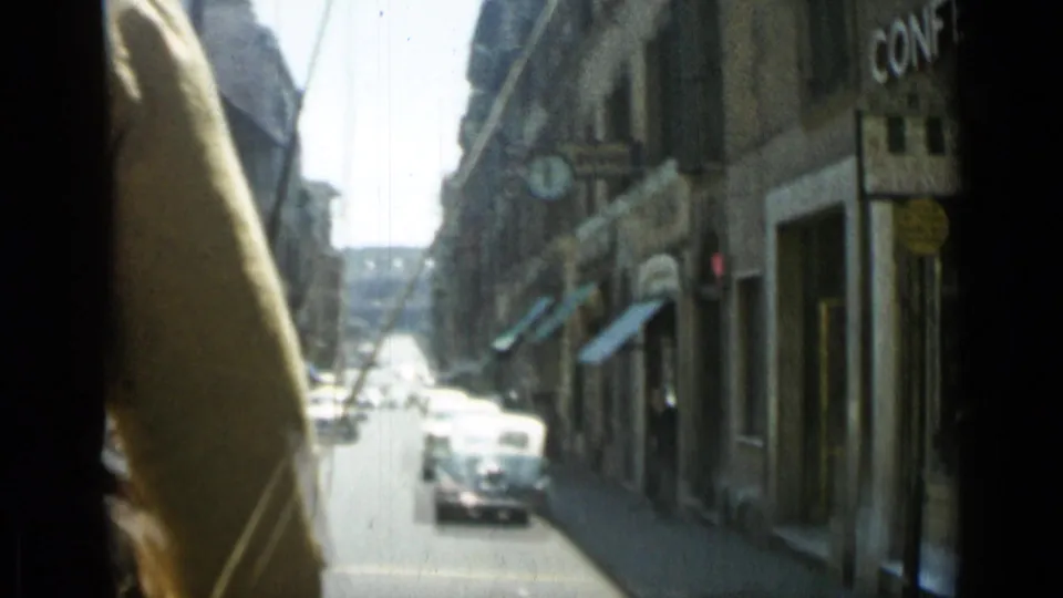 1962 Rome footage from a moving vehicle, with classic cars on a narrow street, shaded shopfronts, stone buildings, and a visible window or frame edge.