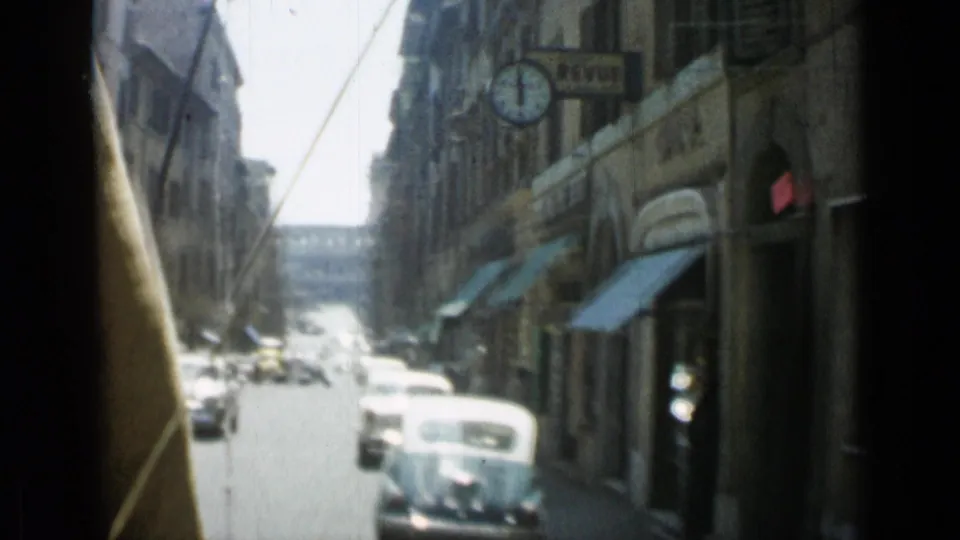 1962 Rome street scene with a classic car in the foreground, storefront awnings, stone facades, signage, and bright traffic farther down the road.