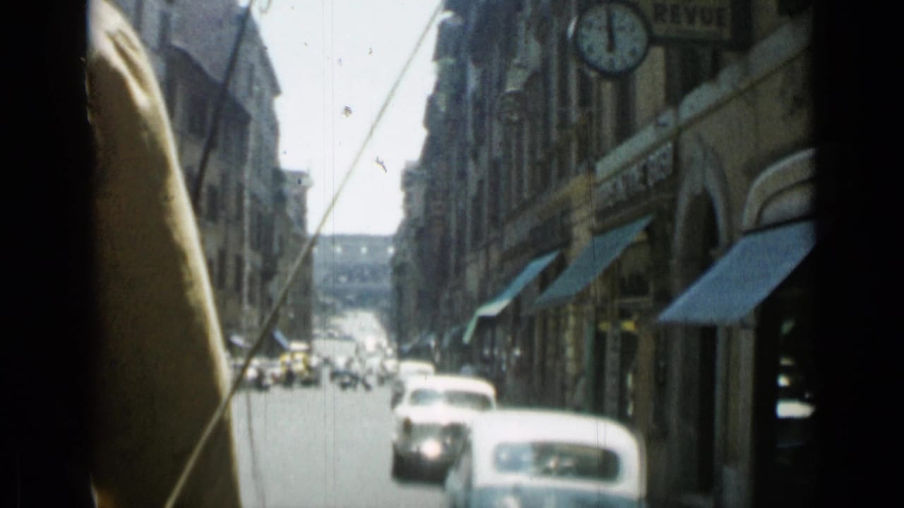 Moving car view down a narrow city street with small cars, tall buildings, awnings, and a round sign or clock.
