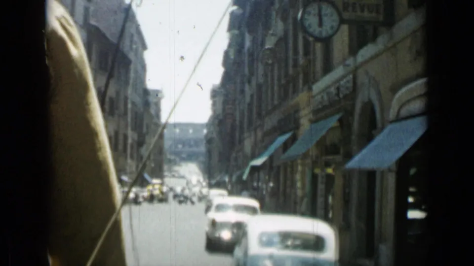 1962 Rome street view from a moving car, showing compact cars, tall stone buildings, shop awnings, and a round street sign or clock.