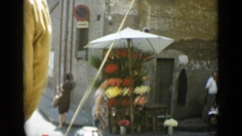 Rome street flower stand under a white umbrella, with pedestrians, stone wall texture, and signs visible in the background.