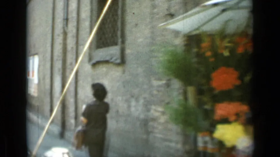 Moving 8mm view in Rome with a pedestrian near a stone wall and a blurred flower display passing close to the camera.