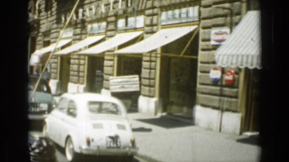 1962 Rome street scene with a white compact car, stone building facades, striped awnings, and shop signs along the sidewalk.