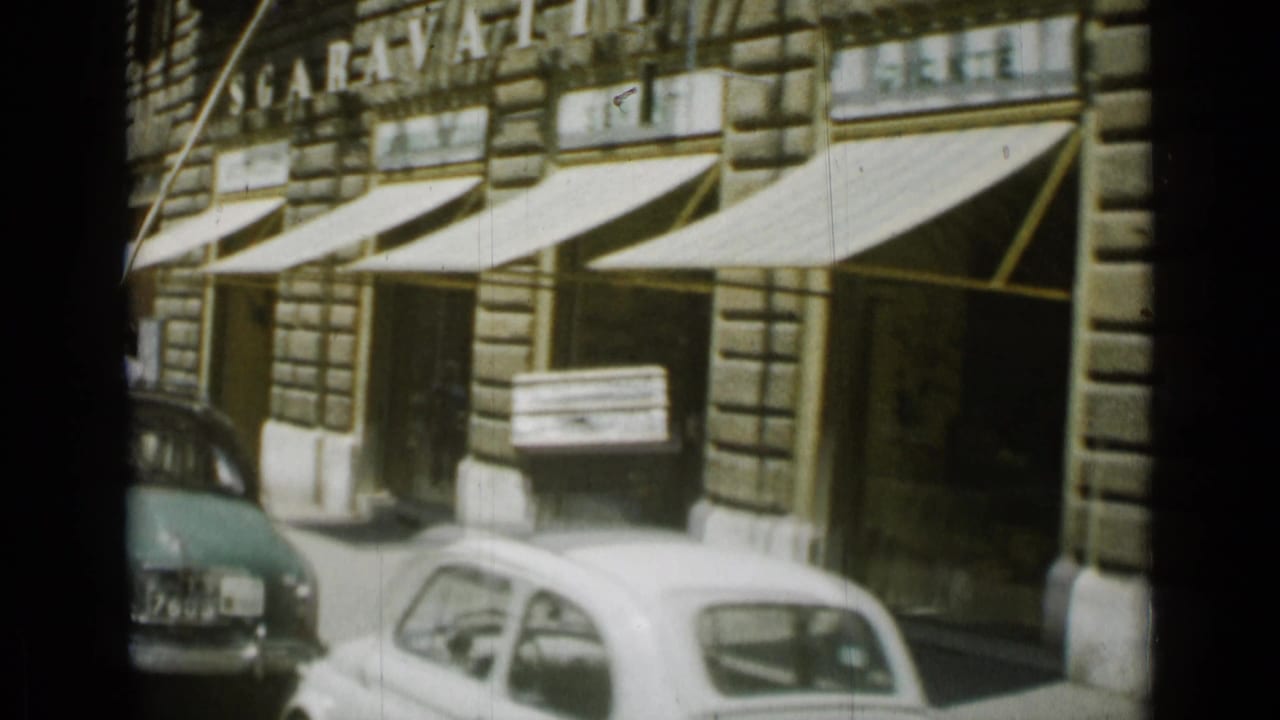 Street storefronts with striped awnings, parked cars, and a man carrying a large crate.