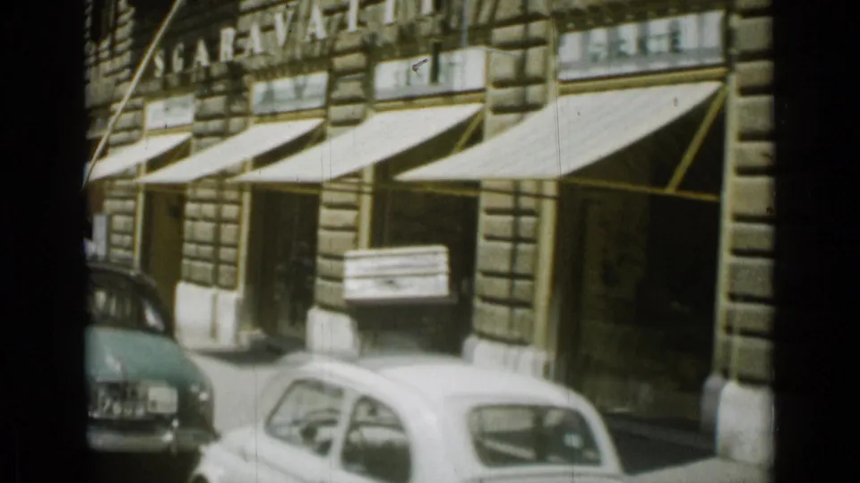 1962 Rome street view with striped awnings, parked small cars, and a man carrying a large crate past shopfronts.