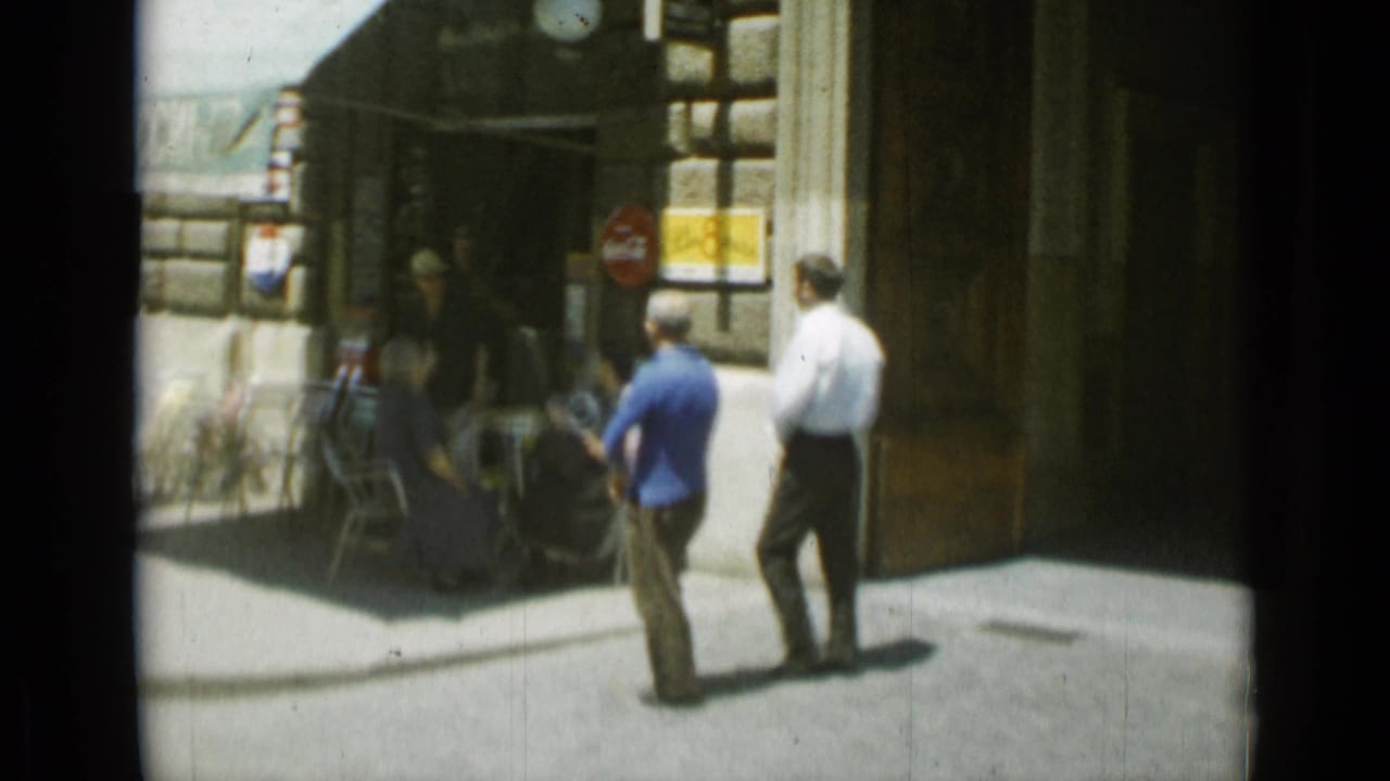 Two men walk near a corner with café seating and a red circular sign.