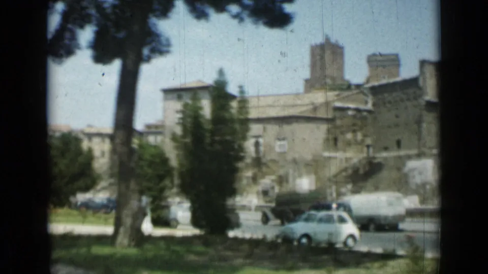 Rome street view with trees in the foreground, small cars at the curb, and large masonry buildings beyond.