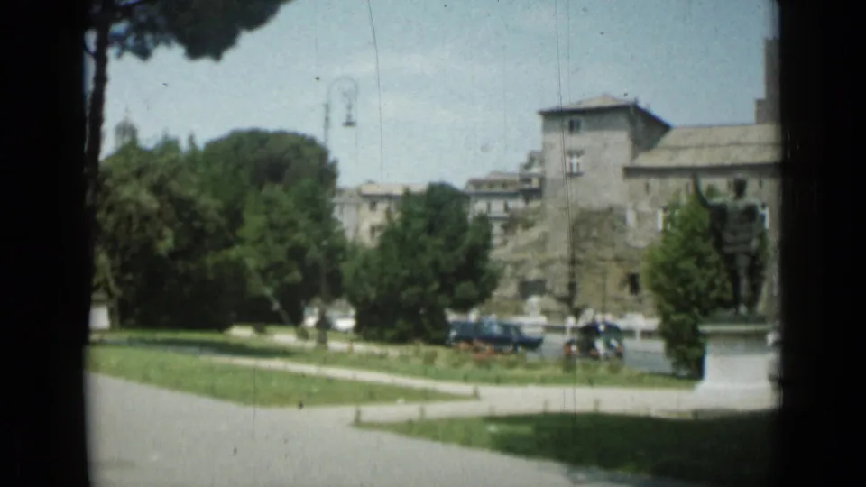 Rome view showing formal lawns, trees, surrounding buildings, mid-century cars, and a statue at the edge of the frame.