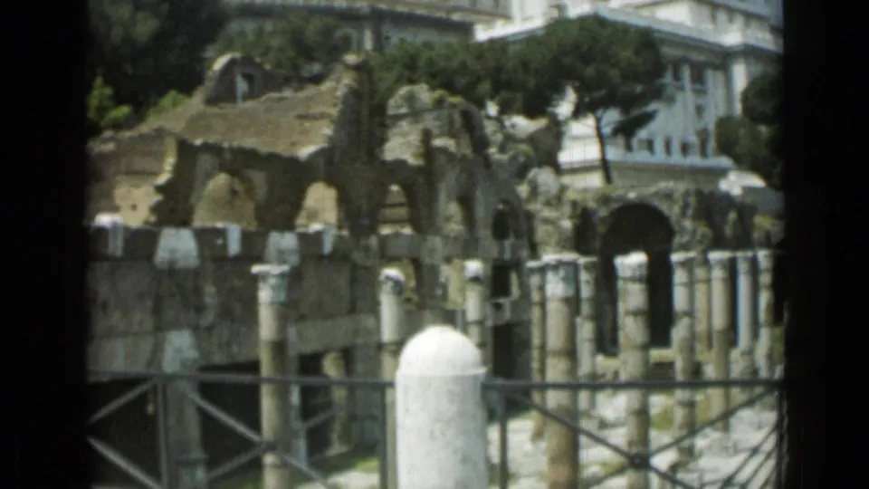 1962 Rome footage of ancient stone ruins with arches and columns, framed by railings, trees, and surrounding buildings.