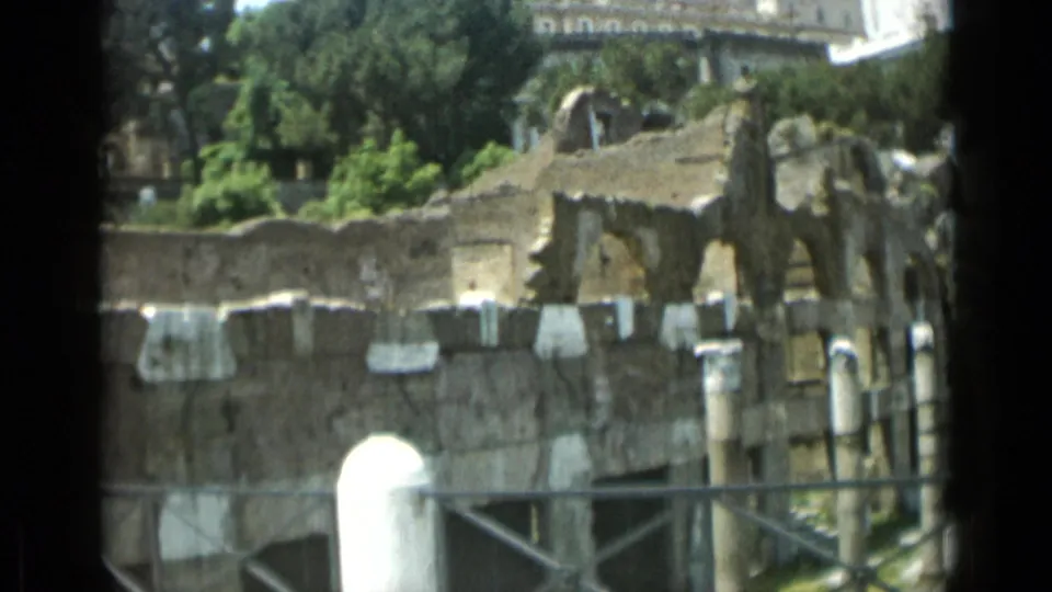 Rome home-movie view of ancient ruins, showing stone arches and columns behind a railing with trees rising behind them.