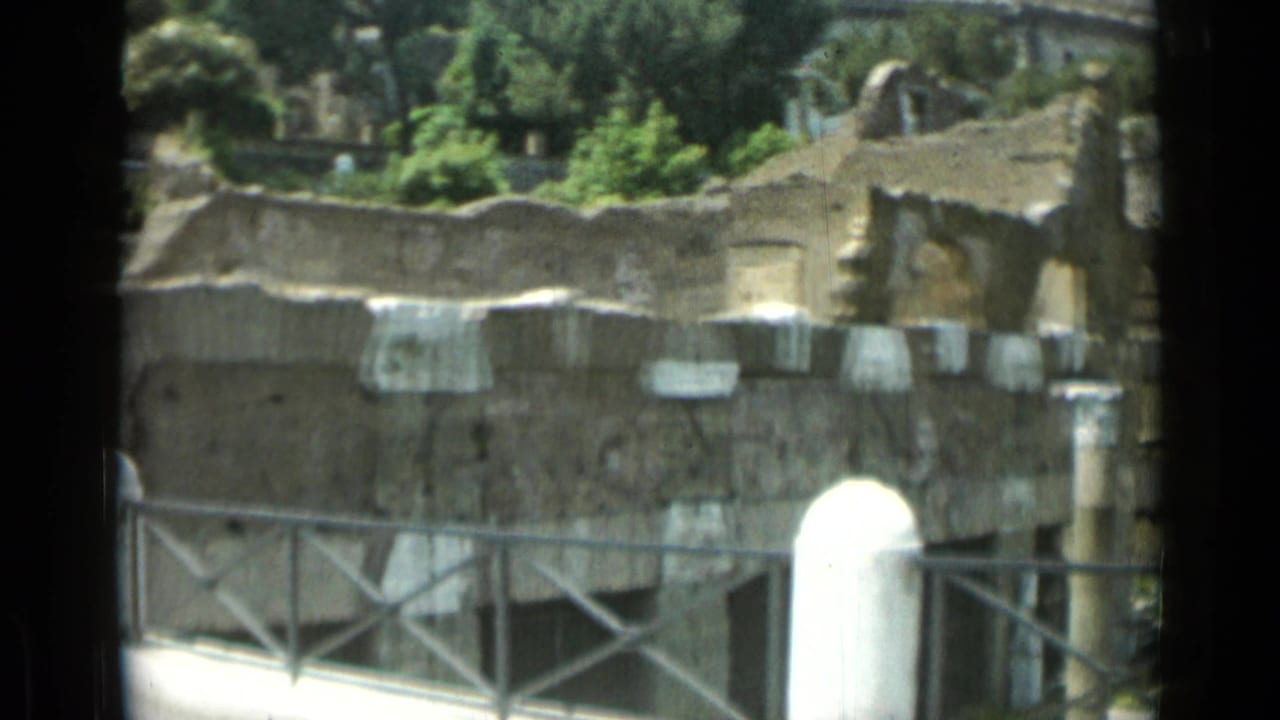 Stone ruins and railings with trees and a white monument partly visible beyond.