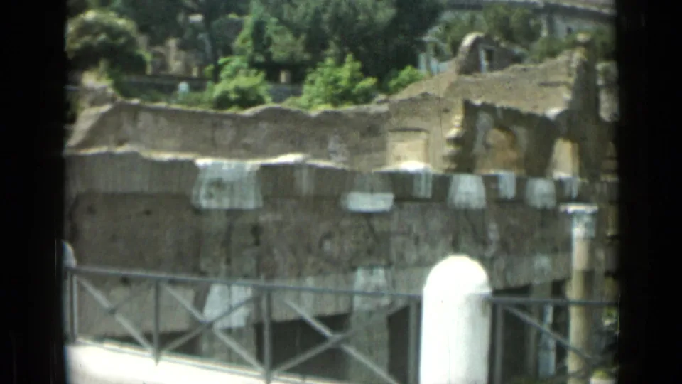 1962 Rome 8mm frame showing weathered stone ruins, metal railings, trees, and part of a white monument in the background.