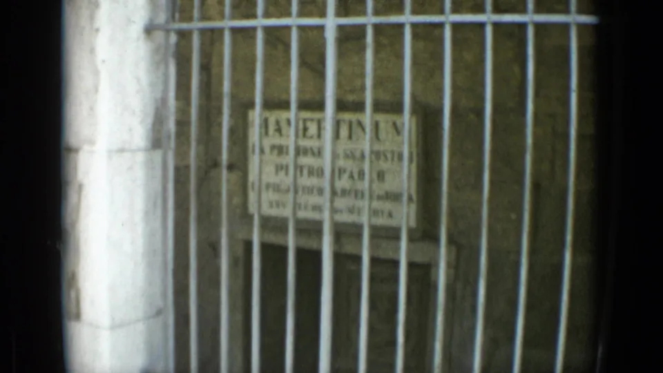 Handheld 1962 Rome travel-film detail of a weathered Latin inscription seen behind vertical bars.