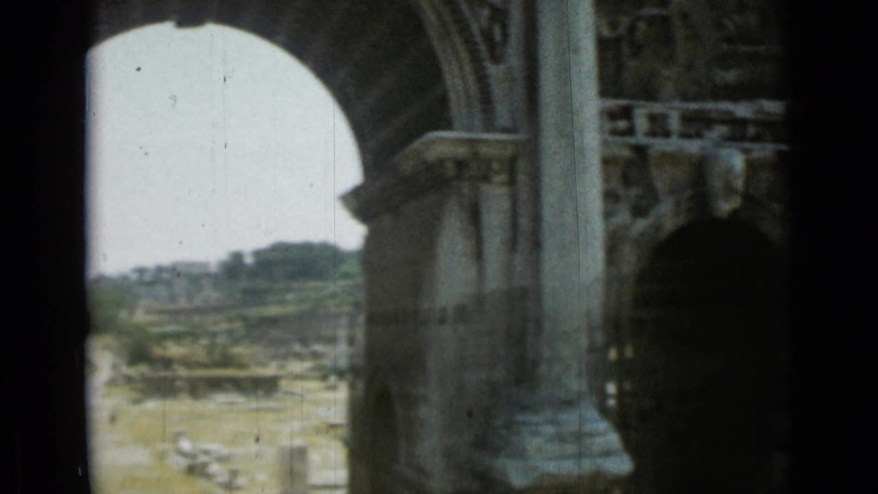 View through a large stone archway in Rome, with carved masonry in the foreground and ancient ruins visible beyond.