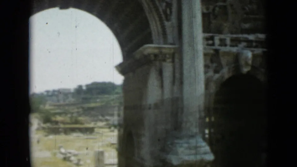 View through a large stone archway in Rome, with carved masonry in the foreground and ancient ruins visible beyond.