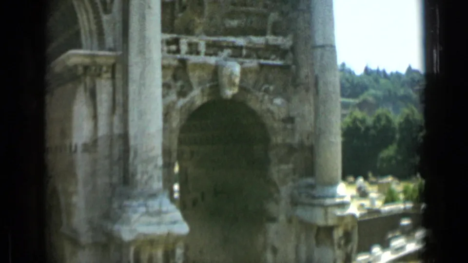 1962 Rome home-movie frame showing a weathered stone arch, column, carved details, and green trees beyond the ruins.