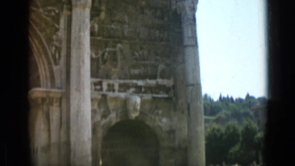 Final 8mm frame from Rome showing a close stone archway, vertical column, carved ruin surfaces, trees, and pale sky.