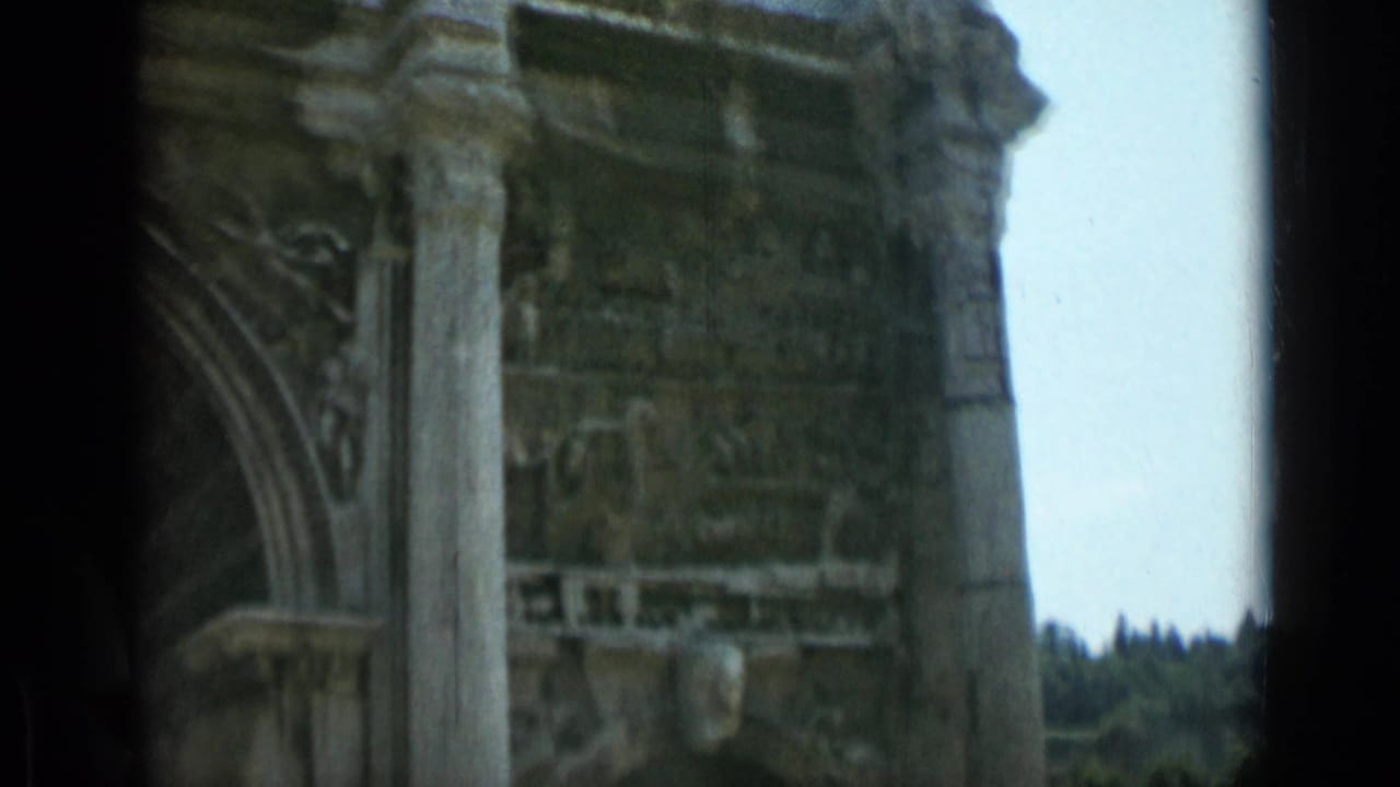 1962 8mm Rome footage showing a close, partial view of the Arch of Constantine's upper stonework, column, carved arch detail, and pale sky.