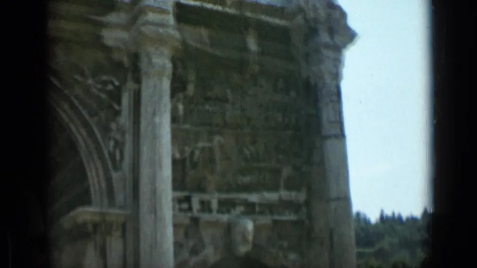 1962 8mm Rome footage showing a close, partial view of the Arch of Constantine's upper stonework, column, carved arch detail, and pale sky.