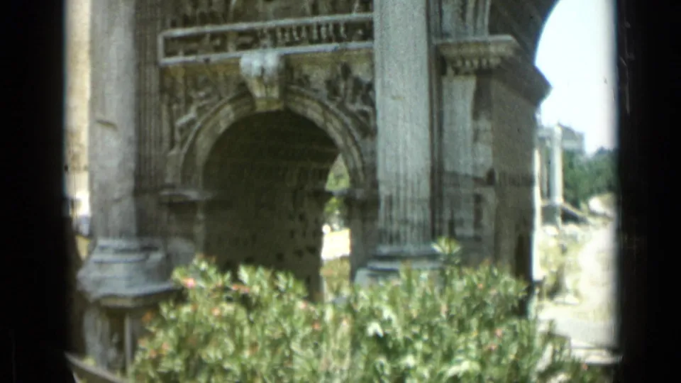 1962 Rome frame showing the monument arch, columns, relief panels, and greenery in the foreground during a handheld pan.